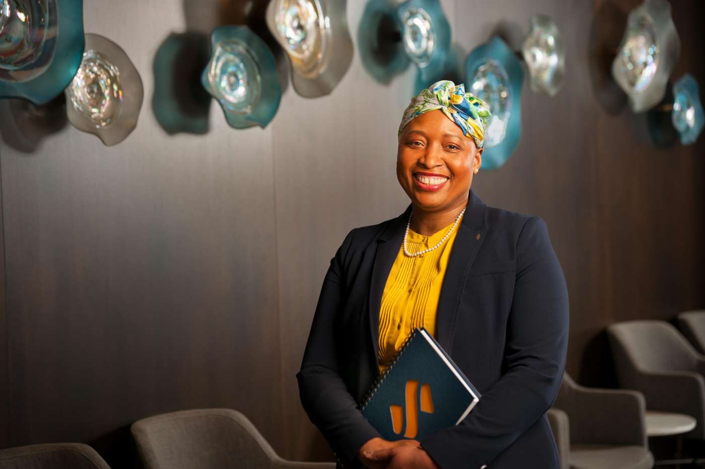 A banking executive poses for a portrait taken with warm lighting in a conference room in front of a wood panel wall adorned with glassy decorative elements.