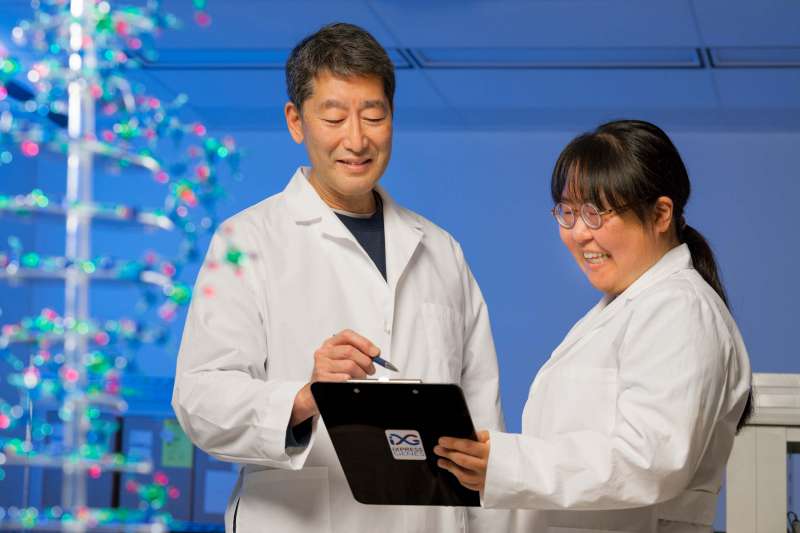 Two scientists in lab coats review findings on a clipboard while standing in a lab with a model of a DNA strand glistening in the foreground.