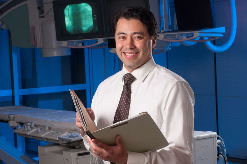 A cardiologist stands in a hospital operating room holding a clipboard.