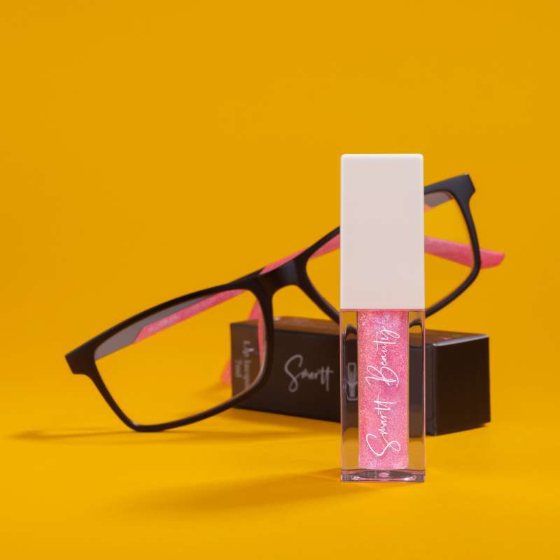 A glassy tube of pink glittery lip gloss photographed with a pair of glasses in this stylized cosmetic product photo.
